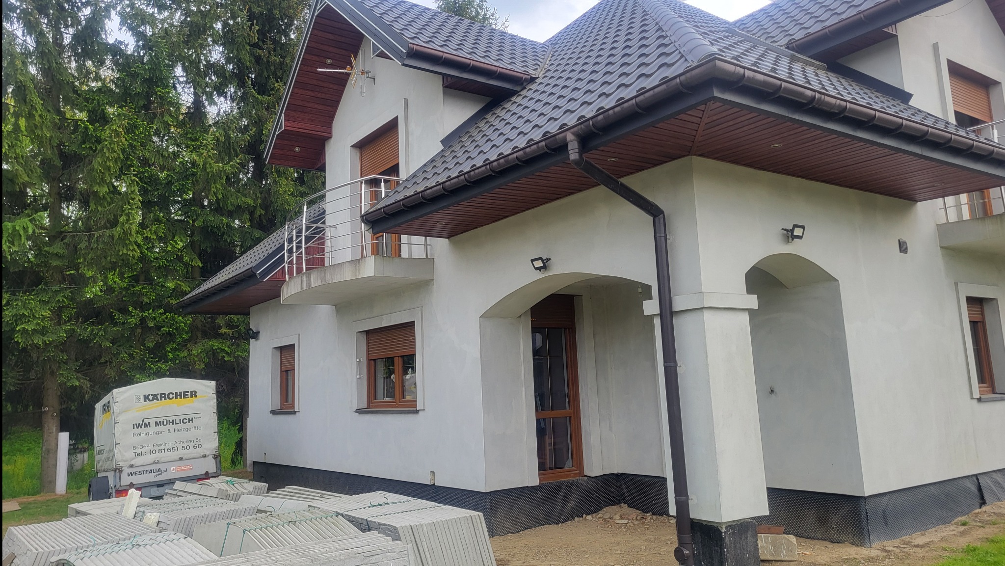 Nowoczesny dom jednorodzinny z balkonem i brązowymi oknami, widok z zewnątrz, z zaparkowaną przyczepą firmy sprzątającej i stertą płyt chodnikowych.