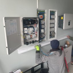 Nexbe - Instalacja elektryczna: elektryk w trakcie montażu rozdzielnicy i podłączenia przewodów wewnątrz budynku. Widoczne narzędzia i schemat.