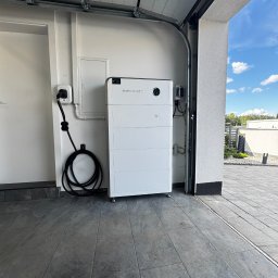 Nexbe - Biały magazyn energii Sideenergy zamontowany w garażu, widoczne okablowanie i gniazdko. Częściowo otwarta brama garażowa, w tle błękitne niebo.