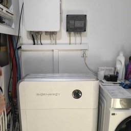 Nexbe - Biały, kompaktowy magazyn energii SIGENERGY w pomieszczeniu gospodarczym, obok pralki. Na ścianie skrzynka elektryczna i moduł sterujący. Widoczne przewody i instalacje.