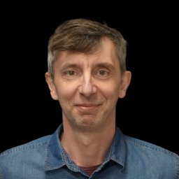 COMMBRIDGE SPÓŁKA Z OGRANICZONĄ ODPOWIEDZIALNOŚCIĄ - Portret uśmiechniętego mężczyzny w dżinsowej koszuli na czarnym tle. Widoczne zmarszczki mimiczne i delikatny zarost. Ujęcie od przodu, skupione na twarzy.