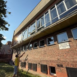 Solex Group sp. z o.o. - Mężczyzna myje okna budynku z cegły za pomocą długiej lancy teleskopowej. Widoczne okna z żaluzjami, trawnik i drzewo. Słoneczny dzień.