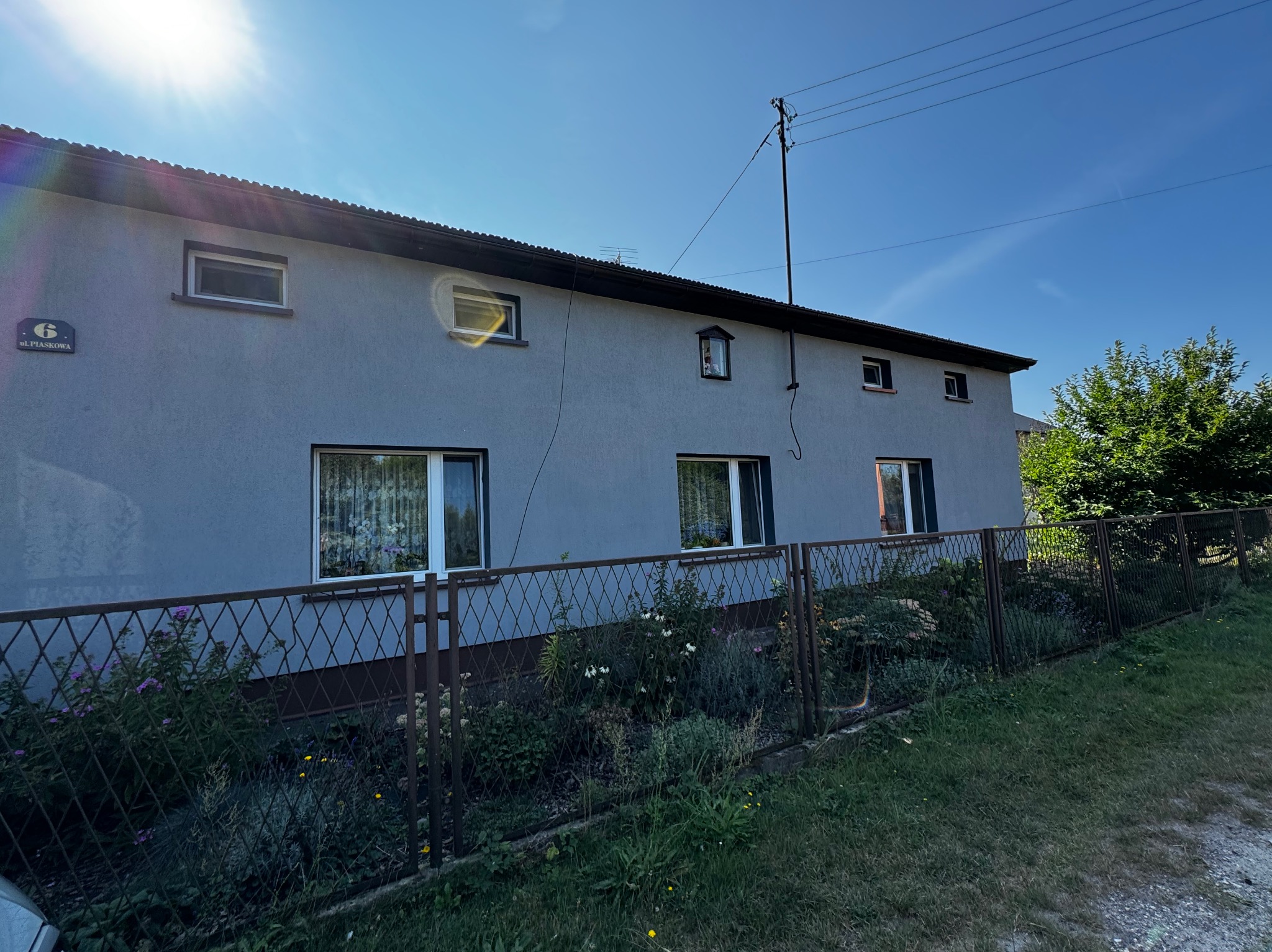 Dwukondygnacyjny dom z szarą elewacją, widoczny z poziomu ogrodu za metalowym ogrodzeniem. Na elewacji widnieje tabliczka z numerem '6 ul. Piaskowa'.