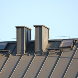 F.H.U TOMBUD - Szary dach z blachy trapezowej z dwoma kominami wentylacyjnymi, dwoma oknami dachowymi i odpowietrznikiem. Widok z dołu na tle jasnego nieba.