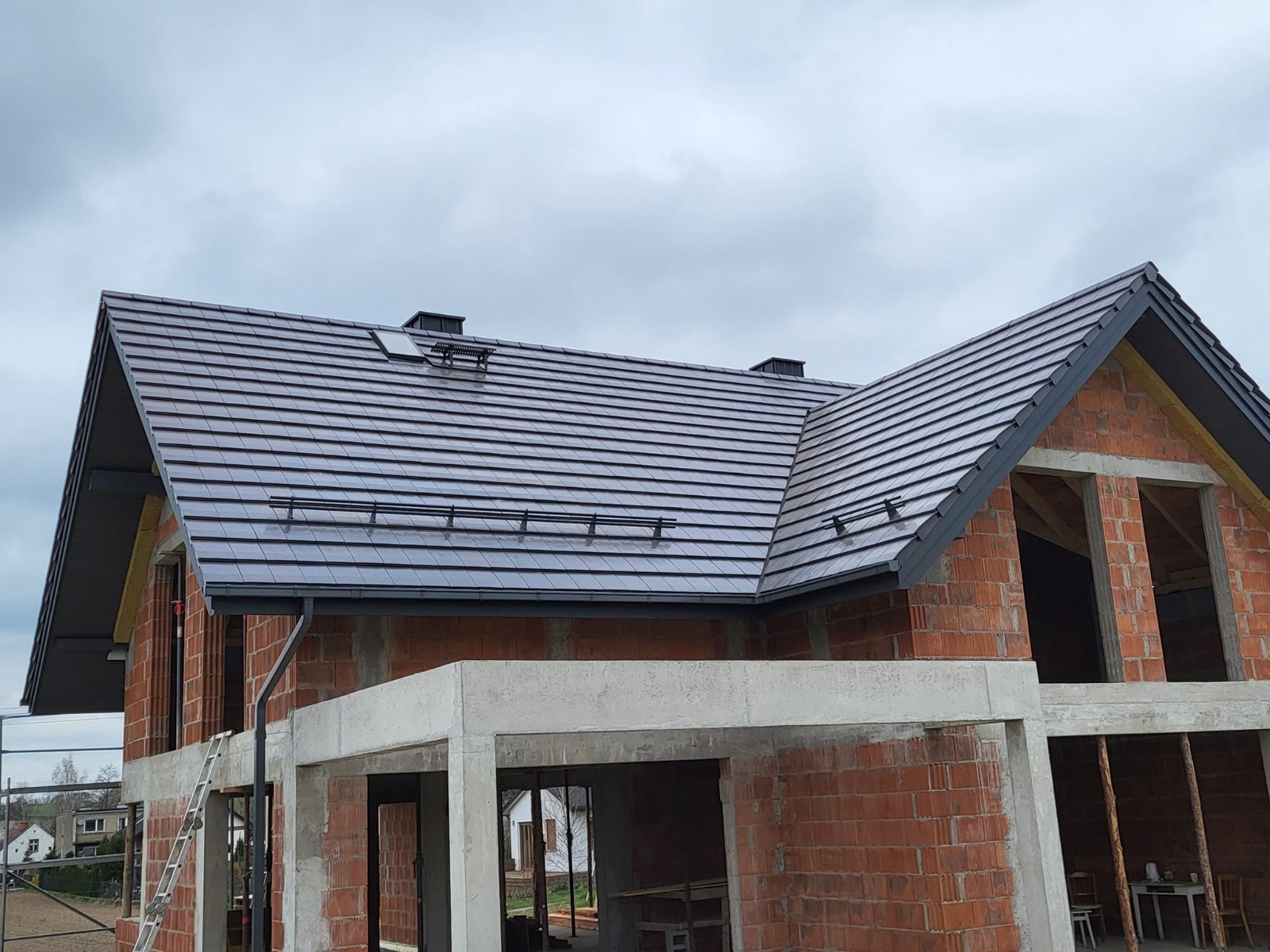 Dom w trakcie budowy z ciemnoszarym, nowym dachem z dachówki ceramicznej. Widoczne kominki wentylacyjne i drabinki przeciwśniegowe. Surowy stan budynku.