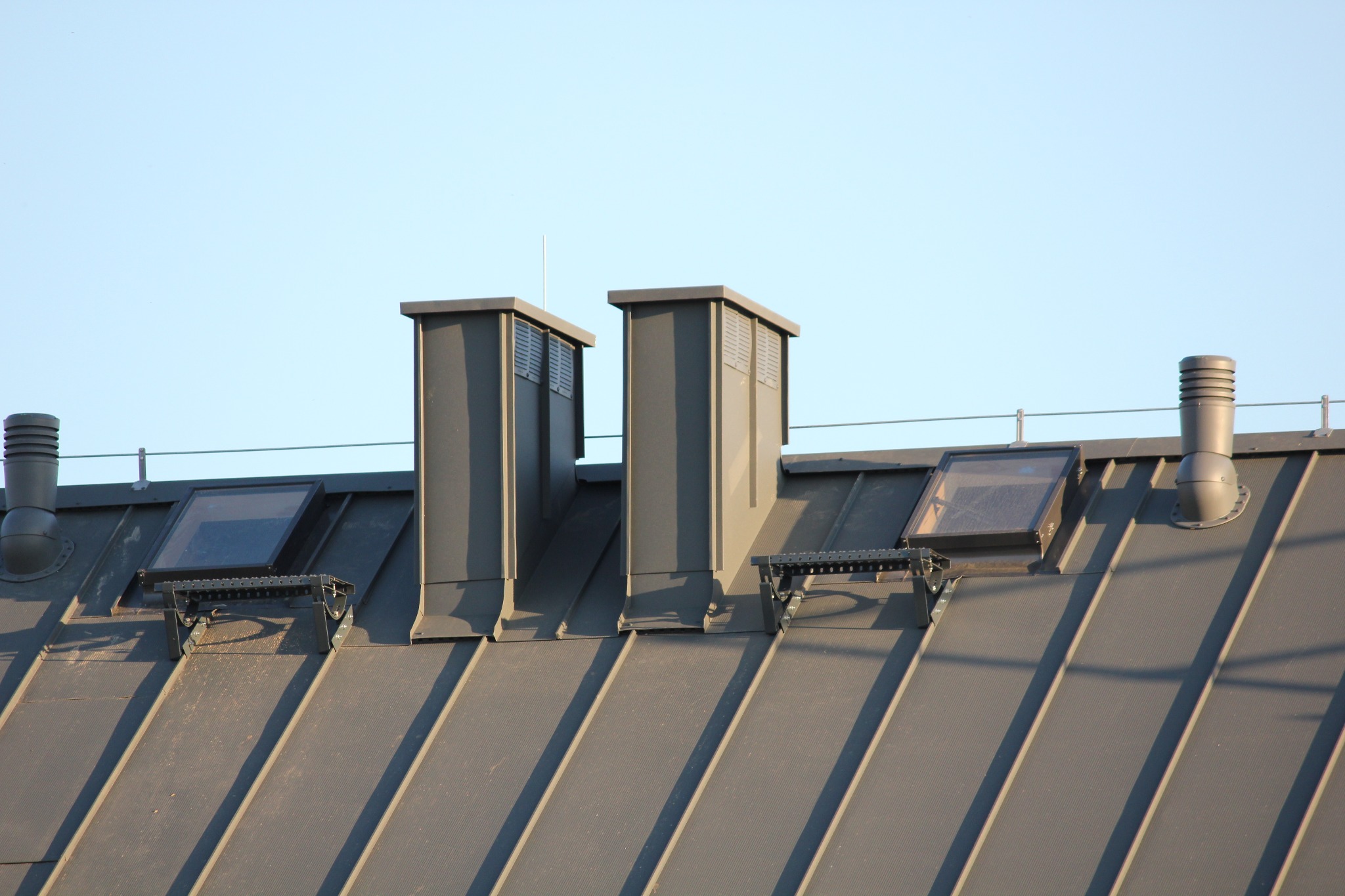 Szary dach z blachy trapezowej z dwoma kominami wentylacyjnymi, dwoma oknami dachowymi i odpowietrznikiem. Widok z dołu na tle jasnego nieba.