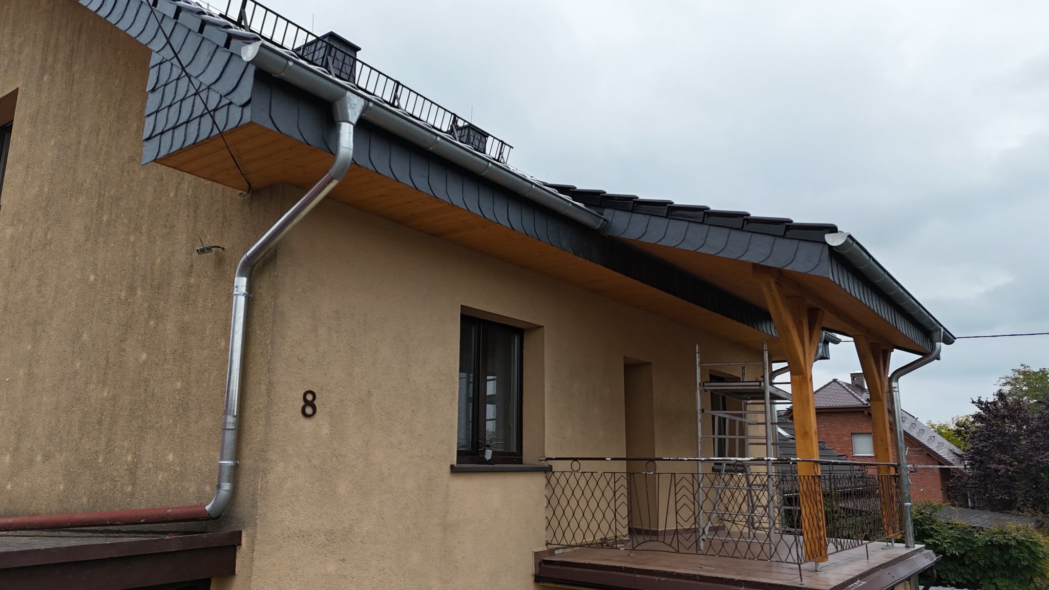 Dom z nowym, grafitowym dachem łupkowym i orynnowaniem. Widoczny fragment balkonu z drewnianą konstrukcją. Na ścianie numer domu '8'.