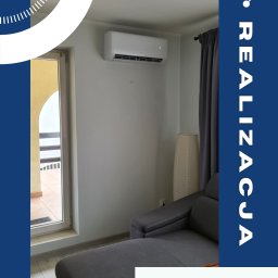 Somar - Biały klimatyzator AUX Q-SMART zamontowany na ścianie w jasnym pokoju z szarą sofą i oknem balkonowym. Widoczna zasłona i lampa podłogowa. Realizacja.