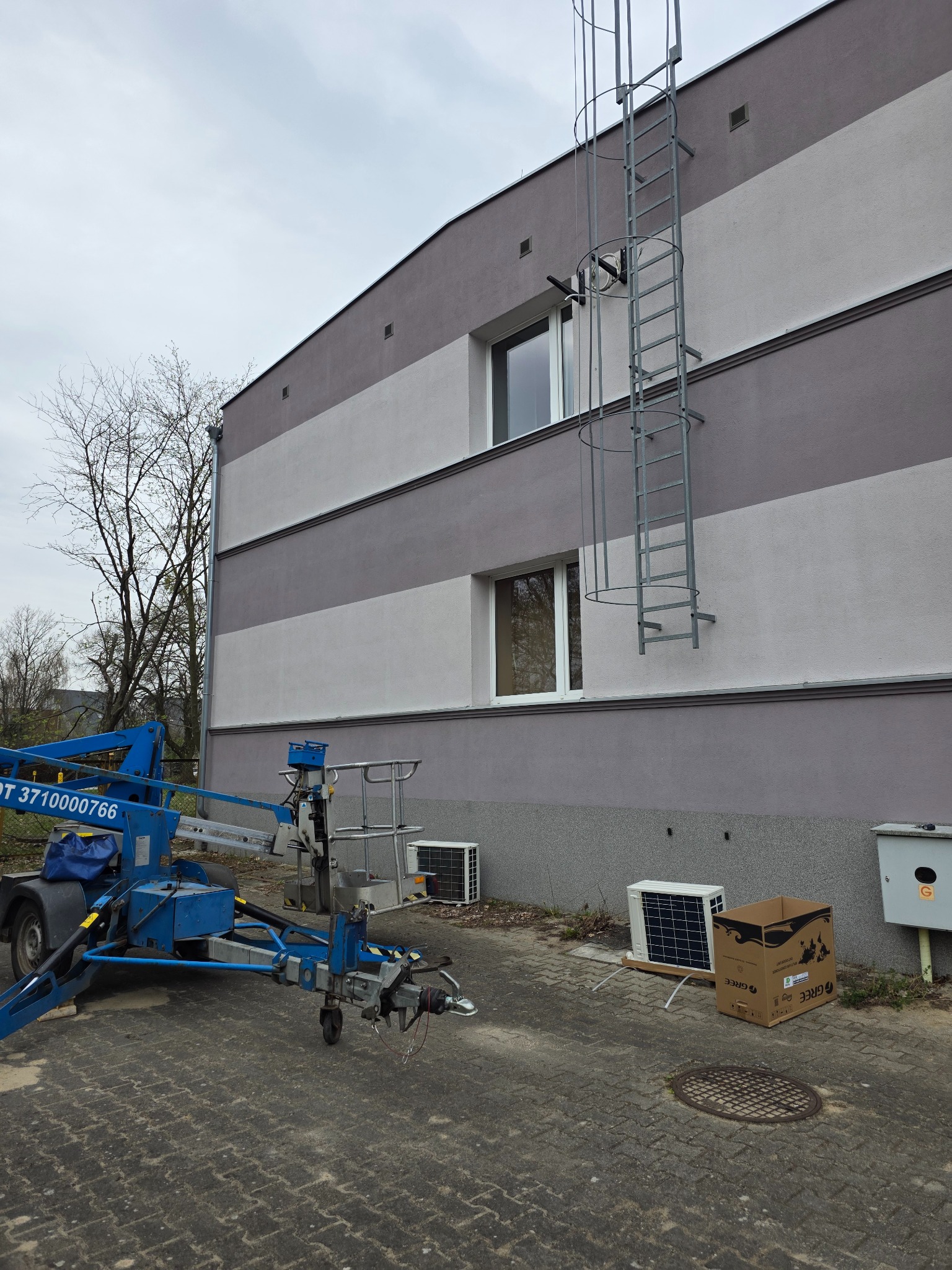 Montaż klimatyzacji zewnętrznej na elewacji budynku z użyciem podnośnika. Widoczna drabina na ścianie i karton po urządzeniu.