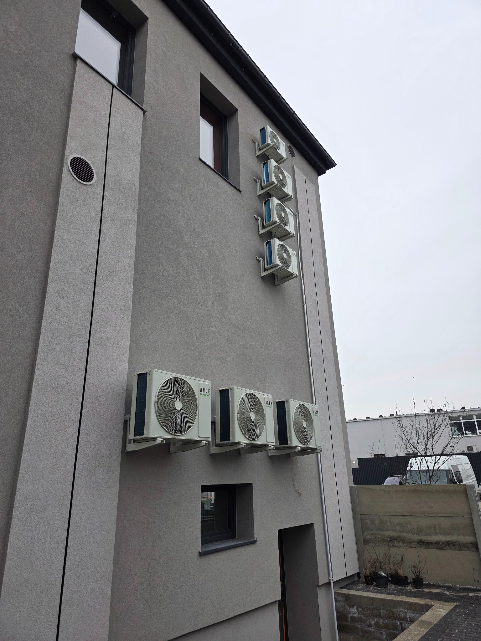 Szereg klimatyzatorów ANDE zamontowanych na szarej elewacji budynku, widoczne jednostki zewnętrzne, rury instalacyjne i fragment otoczenia.