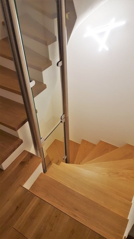 Drewniane schody spiralne z balustradą ze stali nierdzewnej i szkła, oświetlone nowoczesną lampą sufitową. Widok z góry, ukazujący detale wykończenia.