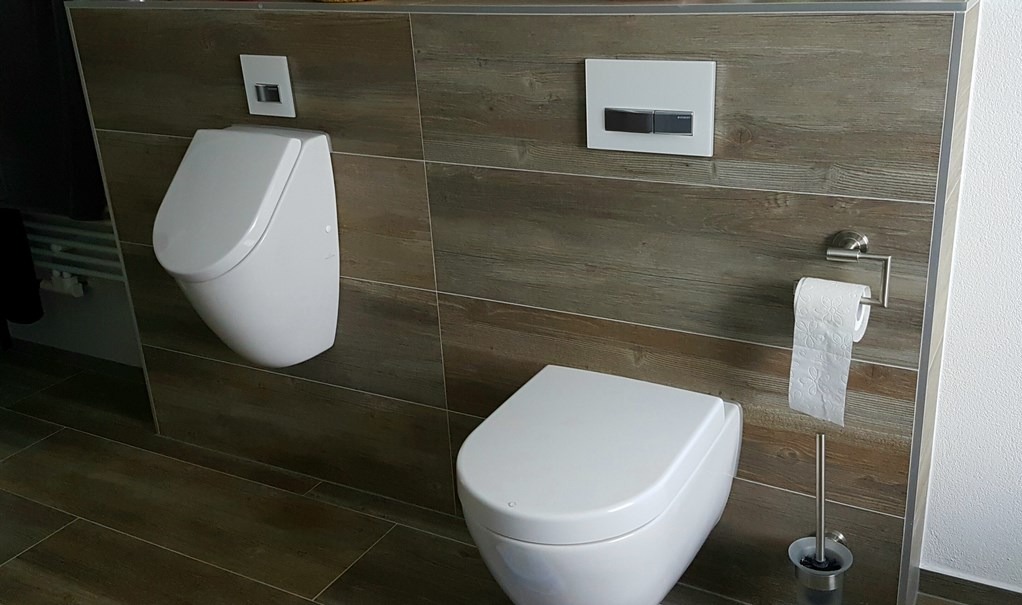 Nowoczesna łazienka z drewnopodobnymi płytkami, wiszącą toaletą i pisuar. Minimalistyczny design, chromowane detale, czystość i funkcjonalność.