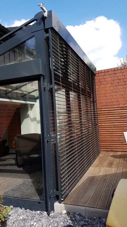 Nowoczesna zabudowa tarasu z szarymi żaluzjami zewnętrznymi, widok z zewnątrz na drewniany taras i ceglany mur. Elementy automatyki na górze konstrukcji.