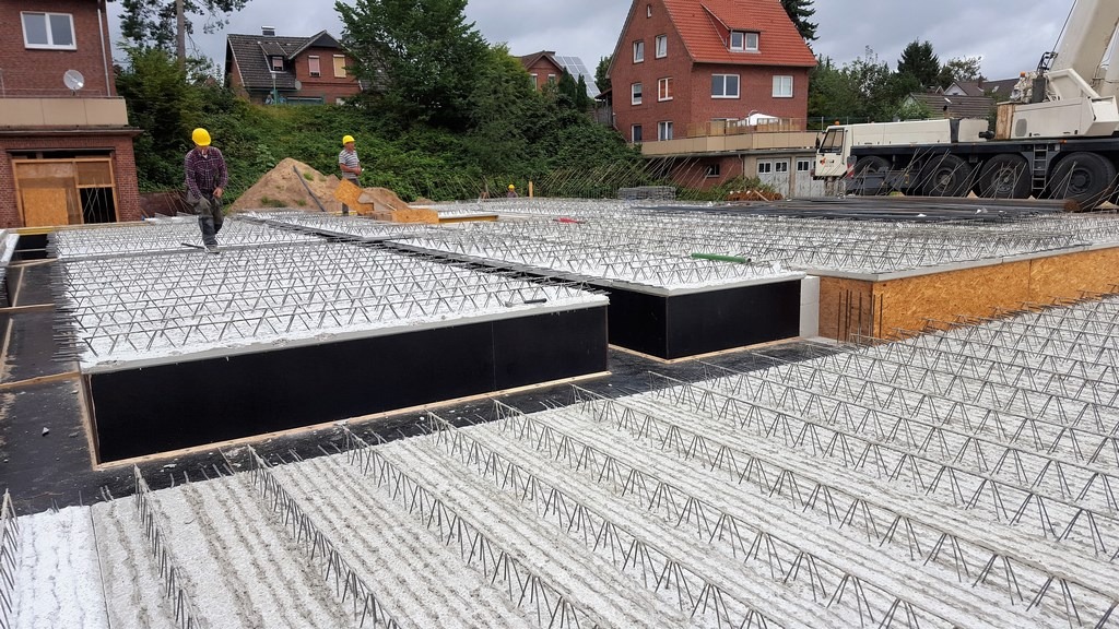 Zbrojenie płyty fundamentowej z pracownikami w żółtych kaskach na budowie domu jednorodzinnego. Widoczny żuraw budowlany i zabudowa osiedlowa w tle.