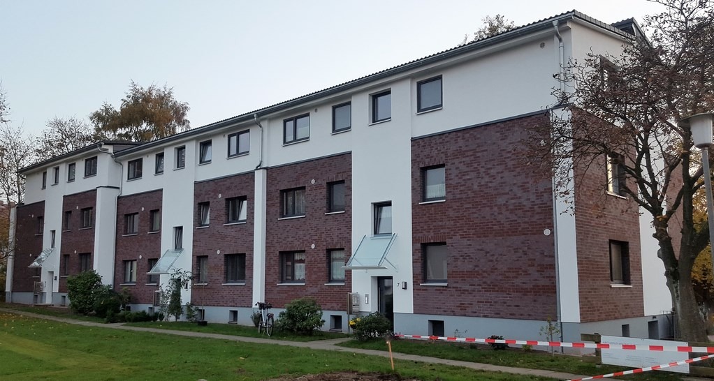 Długi, dwukondygnacyjny budynek z ceglaną elewacją i białymi elementami, widoczny z perspektywy trawnika. Wejścia z daszkami, okna w ciemnych ramach.