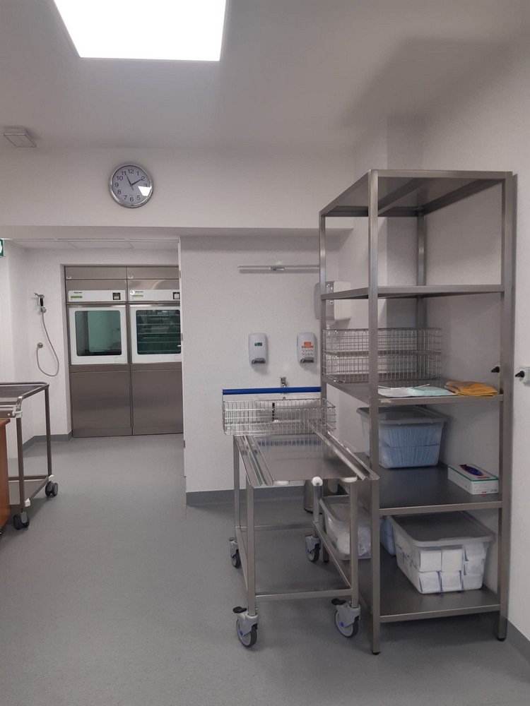 Stalowe regały i wózki w sterylnym pomieszczeniu z szarym linoleum, widoczne umywalki i zegar na ścianie, sugerujące wnętrze placówki medycznej lub laboratorium.