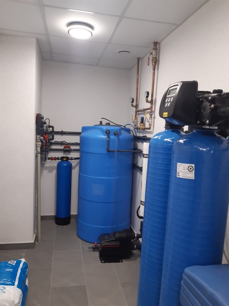 Instalacja uzdatniania wody w pomieszczeniu technicznym w Nawcz. Widoczne niebieskie zbiorniki, rury i armatura. Szare płytki na podłodze, białe ściany i sufit.