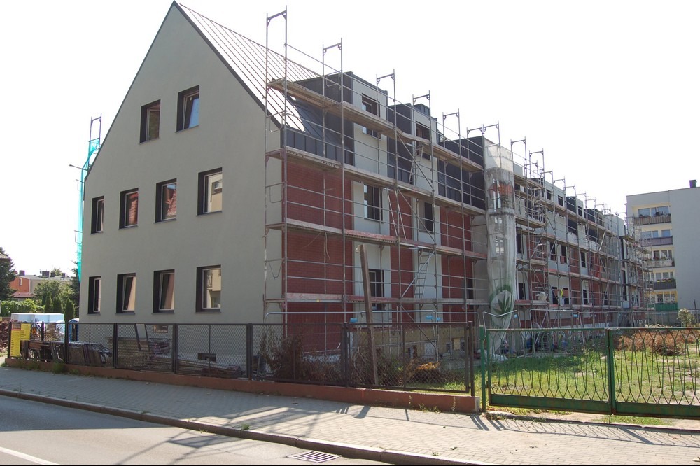 Wielopiętrowy budynek w trakcie ocieplania elewacji, z widocznymi rusztowaniami i fragmentami otynkowanej ściany. Część budynku jest już pomalowana na szaro.