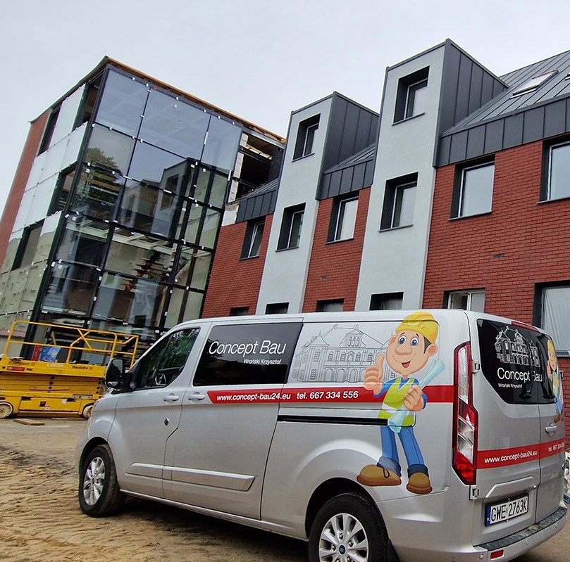 Nowoczesny budynek z przeszkloną fasadą, obok zaparkowany srebrny bus firmy budowlanej z charakterystyczną grafiką i danymi kontaktowymi. W tle widoczny podnośnik.