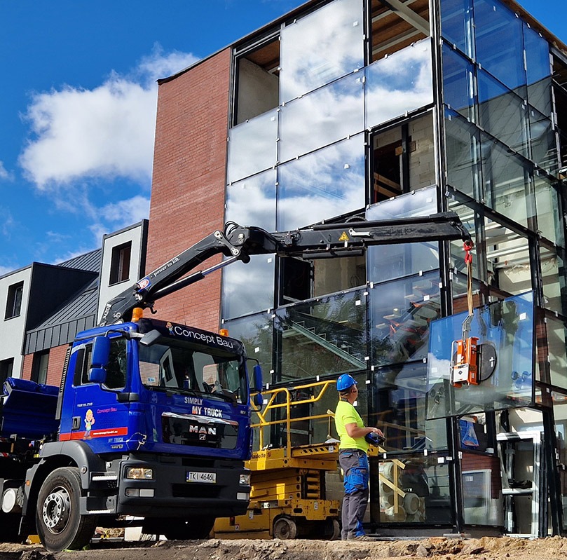 Montaż szklanej fasady budynku z użyciem dźwigu Concept Bau. Pracownik w kasku nadzoruje proces. Widoczna platforma robocza i fragmenty innych budynków w tle.