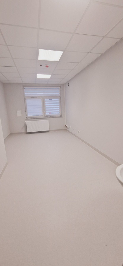 Jasne, puste pomieszczenie po remoncie z grzejnikiem pod oknem z żaluzjami. Widoczne gładkie ściany, podłoga z wykładziny PCV i sufit kasetonowy z oświetleniem.