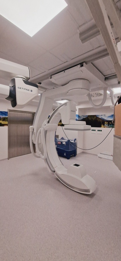 Nowoczesne, jasne wnętrze z aparaturą medyczną Getinge i Philips. Widoczne elementy wyposażenia i wykończenia, w tym podwieszany system sufitowy i grafika na ścianie.