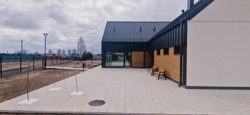 Nowoczesny budynek z ciemnym dachem i drewnianą elewacją, jasny chodnik z kostki, minimalistyczne ławki, surowy krajobraz w tle, pochmurne niebo.