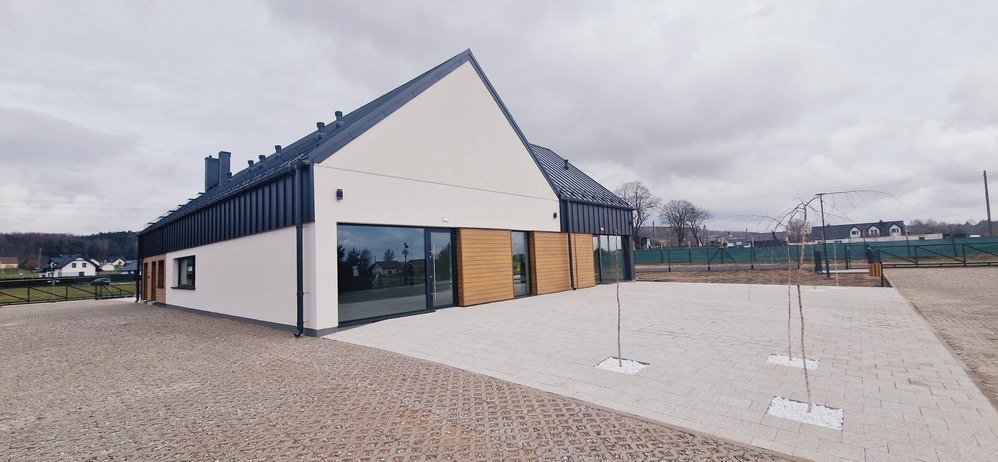 Nowoczesny dom jednorodzinny z ciemnym dachem i jasną elewacją, z elementami drewnianymi. Widoczne duże okna i zagospodarowany teren wokół budynku. Pochmurne niebo w tle.