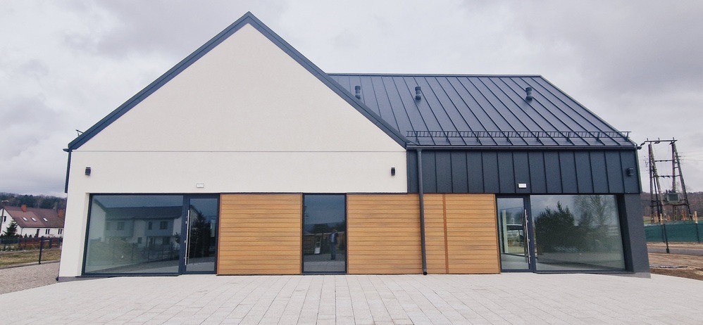 Nowoczesny dom z jasną elewacją i ciemnym dachem, drewniane panele między oknami, widok z zewnątrz na tle pochmurnego nieba i krajobrazu.