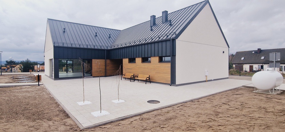 Nowoczesny dom z ciemną, metalową dachówką i jasną elewacją, drewnianymi akcentami, widok z zewnątrz na dziedziniec z ławkami i młodymi drzewkami.