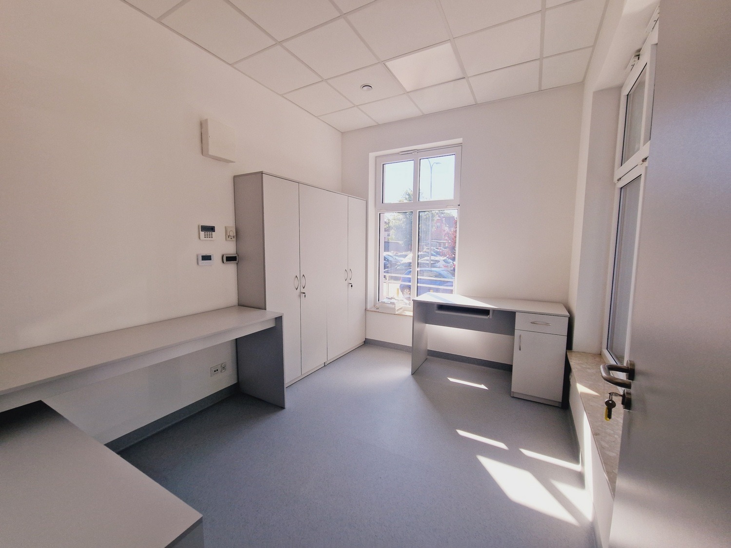 Jasne wnętrze biurowe z szarymi meblami: biurka wzdłuż ścian, szafy zamykane na klucz, okno z widokiem na zewnątrz. Minimalistyczny design, świeżo po remoncie.