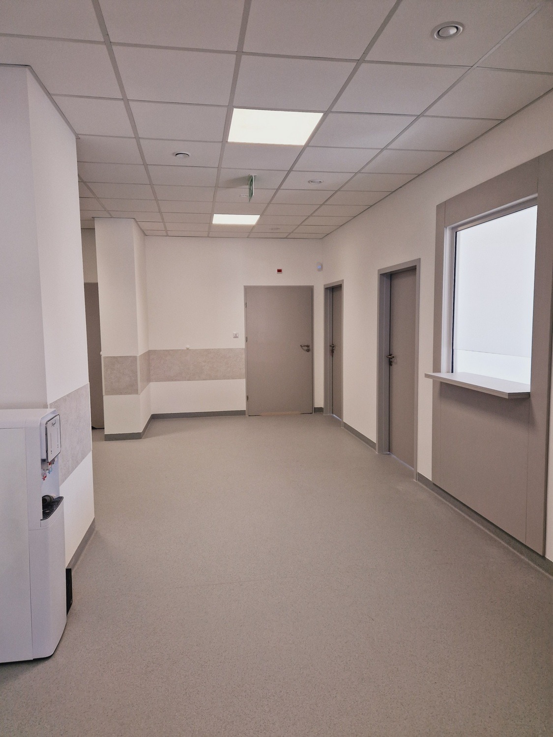 Korytarz w neutralnych barwach z podwieszanym sufitem i szarymi drzwiami. Widoczny dystrybutor wody i okienko podawcze. Ukończone wykończenie wnętrza.