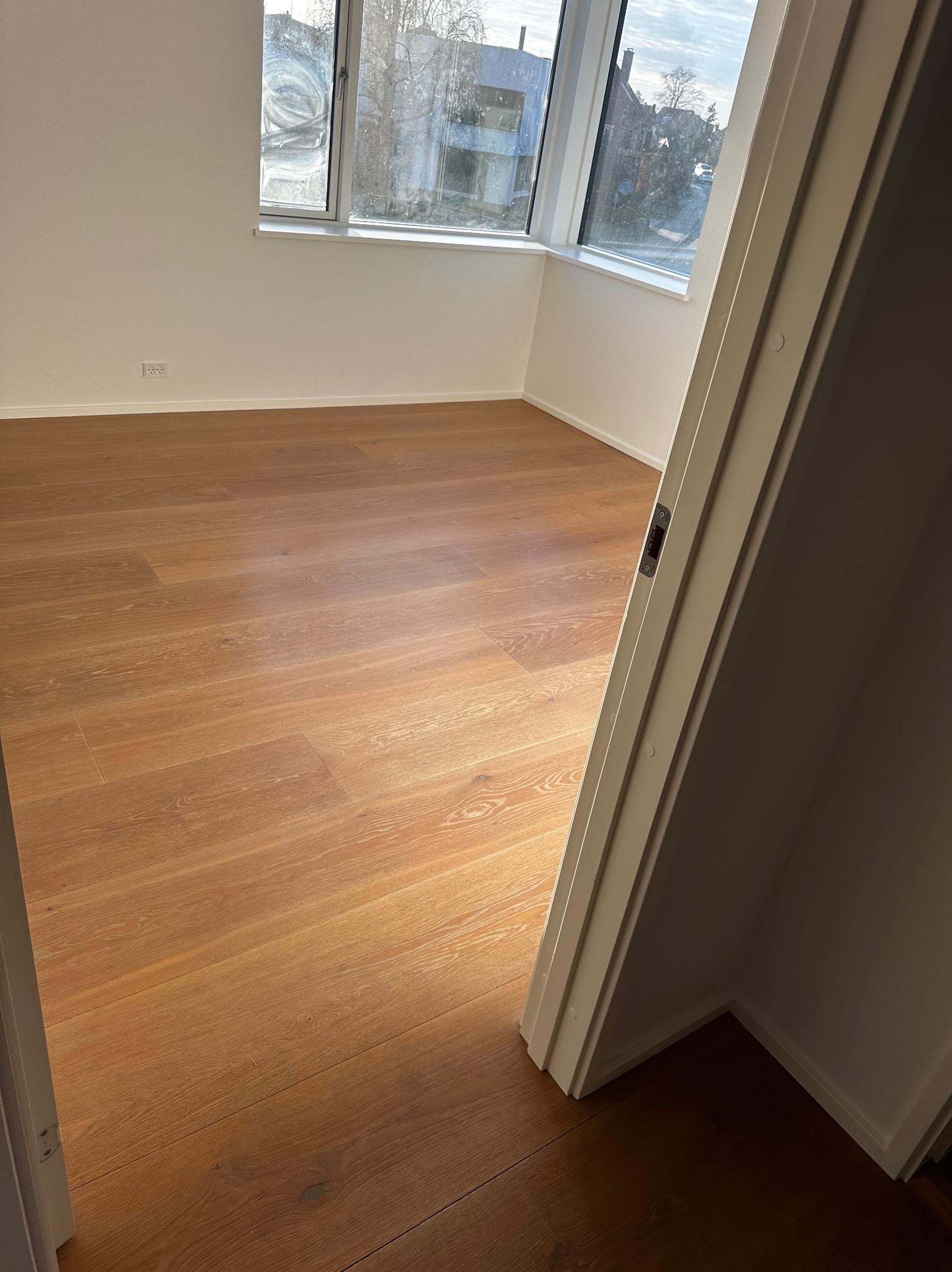 Jasne, puste wnętrze po malowaniu z drewnianą podłogą i widokiem z okna, uchwycone zza otwartych drzwi. Świeżo odmalowane ściany i czysta przestrzeń.