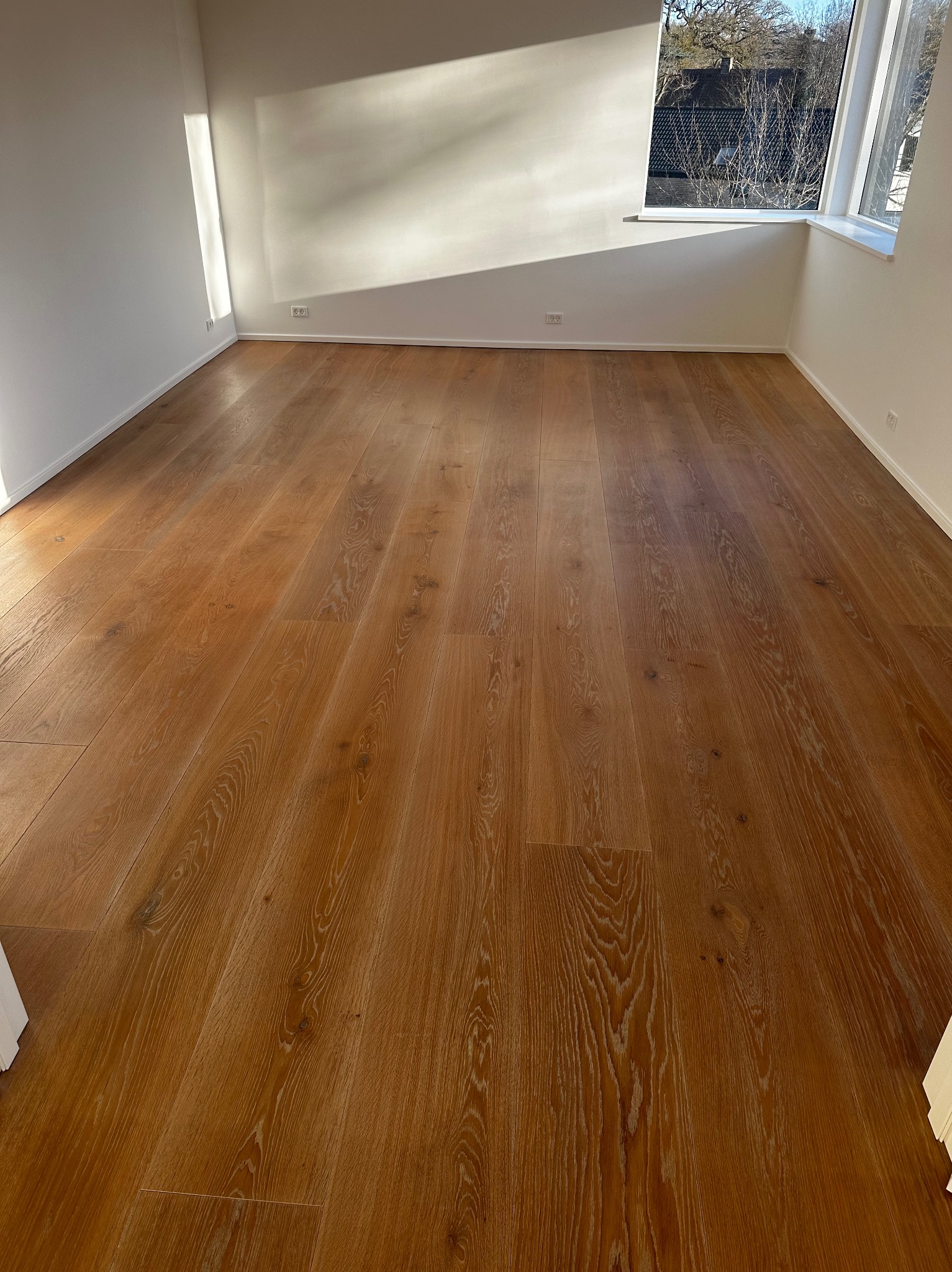 Jasne, drewniane panele podłogowe w pustym pokoju z białymi ścianami i oknem w rogu, wpadające światło słoneczne. Widok na zewnątrz przez okno.