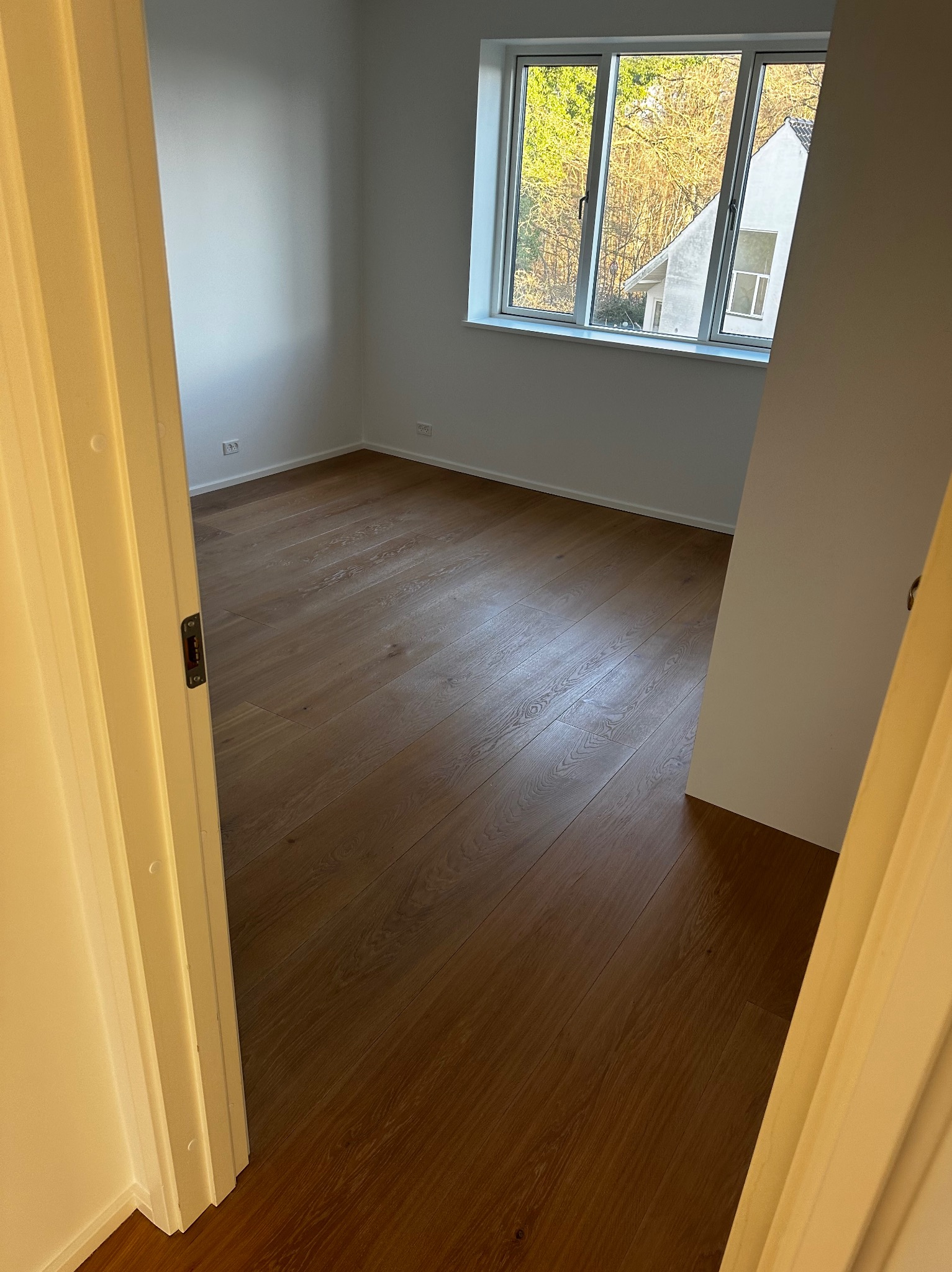 Jasne, puste pomieszczenie z drewnianą podłogą, widok zza otwartych drzwi. Okno z widokiem na drzewa i budynek w tle. Minimalistyczne wnętrze po remoncie.