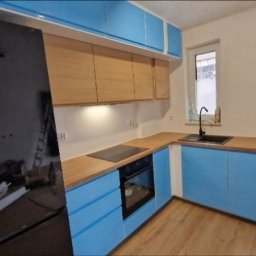 grzegorz skarzynski - Niebiesko-drewniana kuchnia po remoncie: błyszczące fronty szafek, drewniany blat, czarny zlew i płyta grzewcza. Widok na okno i częściowo zasłonięte drzwi balkonowe.