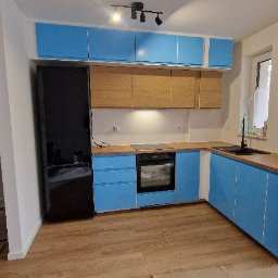 grzegorz skarzynski - Nowoczesna kuchnia z błękitnymi szafkami i drewnianym blatem. Widoczny czarny sprzęt AGD i jasna podłoga. Świeżo po remoncie, folia ochronna na drzwiach w tle.