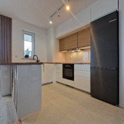 grzegorz skarzynski - Nowoczesna kuchnia z wyspą: białe szafki, drewniane akcenty, czarna armatura i lodówka. Minimalistyczny design, jasne oświetlenie, parkiet w neutralnym odcieniu.