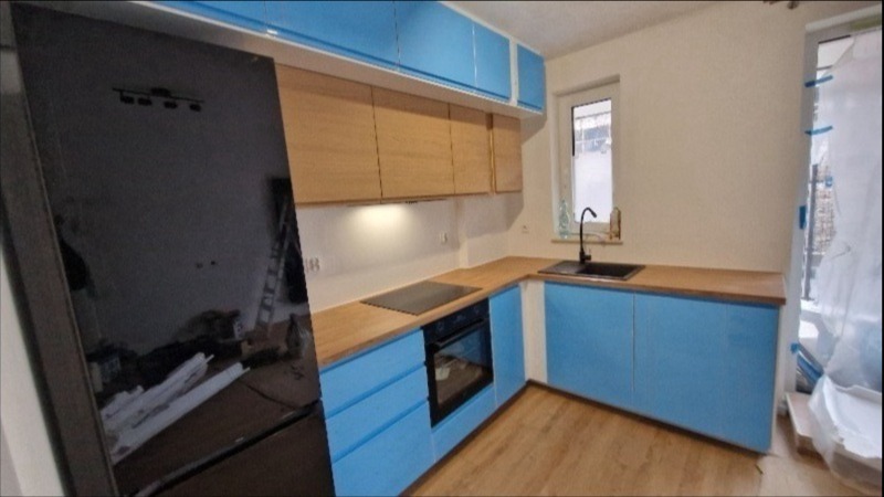 Niebiesko-drewniana kuchnia po remoncie: błyszczące fronty szafek, drewniany blat, czarny zlew i płyta grzewcza. Widok na okno i częściowo zasłonięte drzwi balkonowe.