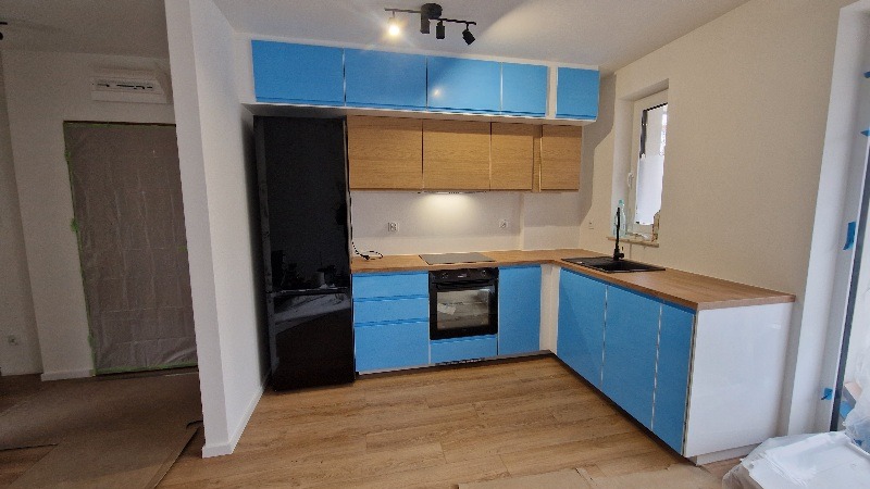 Nowoczesna kuchnia z błękitnymi szafkami i drewnianym blatem. Widoczny czarny sprzęt AGD i jasna podłoga. Świeżo po remoncie, folia ochronna na drzwiach w tle.