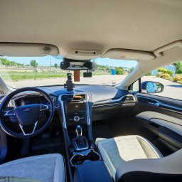 Travel Comfort Kielce - Wnętrze Forda Fusion z jasną tapicerką, widok z tylnego siedzenia na deskę rozdzielczą, kierownicę i przednią szybę z widokiem na parking i zieleń.