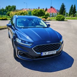 Travel Comfort Kielce - Błyszczący, granatowy Ford Mondeo kombi, zaparkowany na placu, z widoczną tablicą rejestracyjną TK 291EY i budynkiem w tle. Słoneczny dzień.