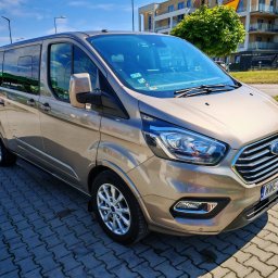 Travel Comfort Kielce - Beżowy Ford Transit Custom z chromowanymi detalami, zaparkowany na kostce brukowej. W tle budynek mieszkalny i stacja paliw.