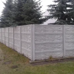 Długi, szary mur z elementów o fakturze kamienia, ciągnący się wzdłuż drogi, z wysokimi świerkami w tle.