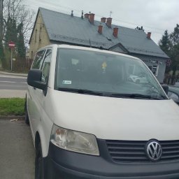 SR transport - Biały Volkswagen Transporter T5 na tle budynku z szarym dachem i szyldem 'Doggy Dog'. Widoczny fragment ulicy i drzewa. Tablica rejestracyjna z przodu pojazdu.