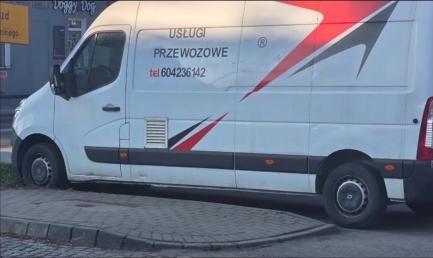 Biały bus z napisem 'Usługi Przewozowe' i numerem telefonu, zaparkowany na chodniku. Widoczny bok pojazdu z elementami graficznymi w kolorze czerwonym i czarnym.