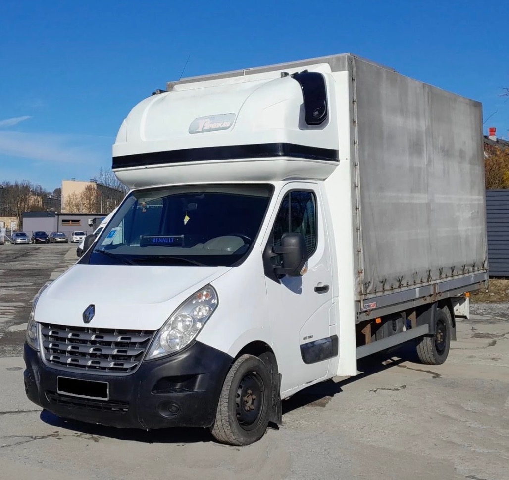 Biały Renault Master z plandeką, wysoki dach, widok z przodu i boku na tle parkingu i błękitnego nieba. Detal: czarne felgi i osłona chłodnicy.