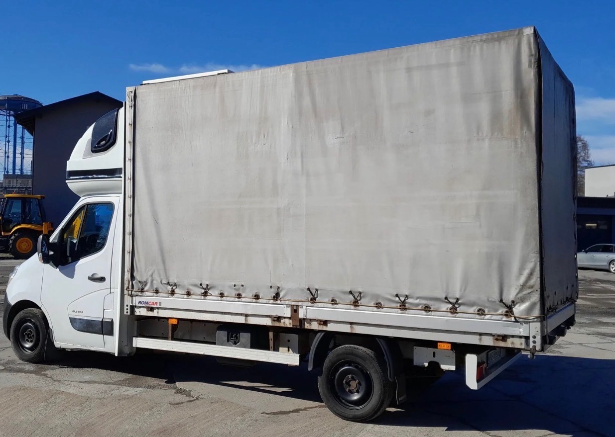 Biały samochód dostawczy z plandeką, widok z boku na tle błękitnego nieba i zabudowań. Na plandece widoczne ślady użytkowania. Z tyłu zaparkowany srebrny samochód.