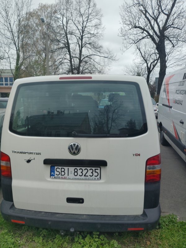 Tył białego busa VW Transporter TDI z naklejką helikoptera i numerem rejestracyjnym SBI 83235 na tle drzew i innego busa z usługami przewozowymi.