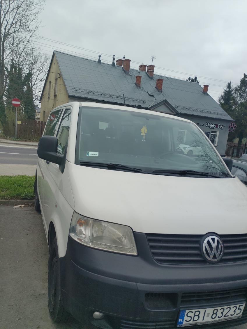 Biały Volkswagen Transporter T5 na tle budynku z szarym dachem i szyldem 'Doggy Dog'. Widoczny fragment ulicy i drzewa. Tablica rejestracyjna z przodu pojazdu.
